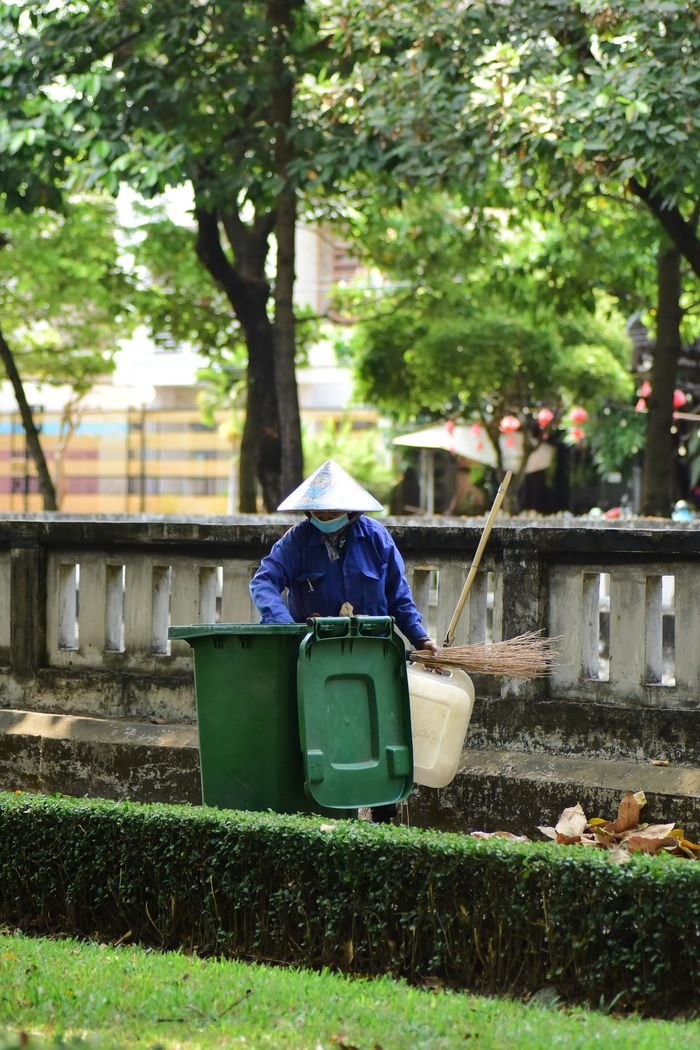 A park worker wears a conical hat while maintaining a clean environment with broom and bin.